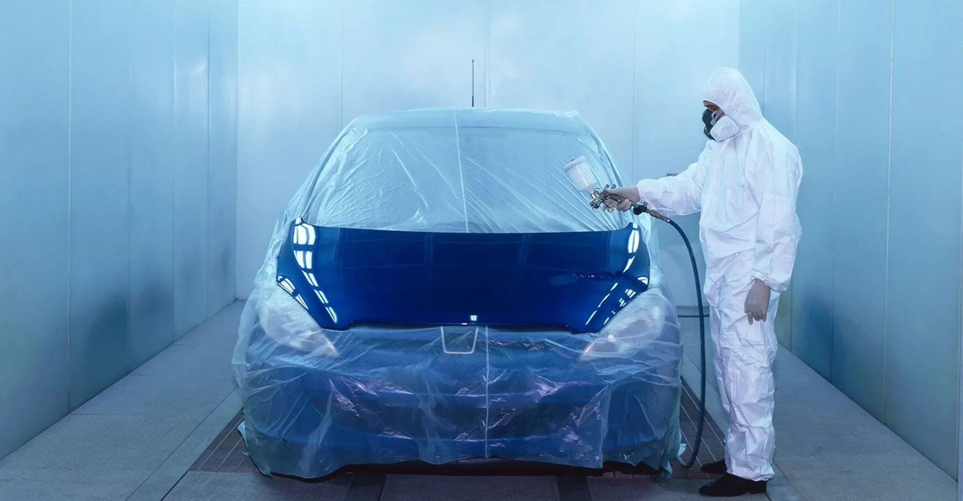 Painter in a white suit spraying a masked car inside a paint booth